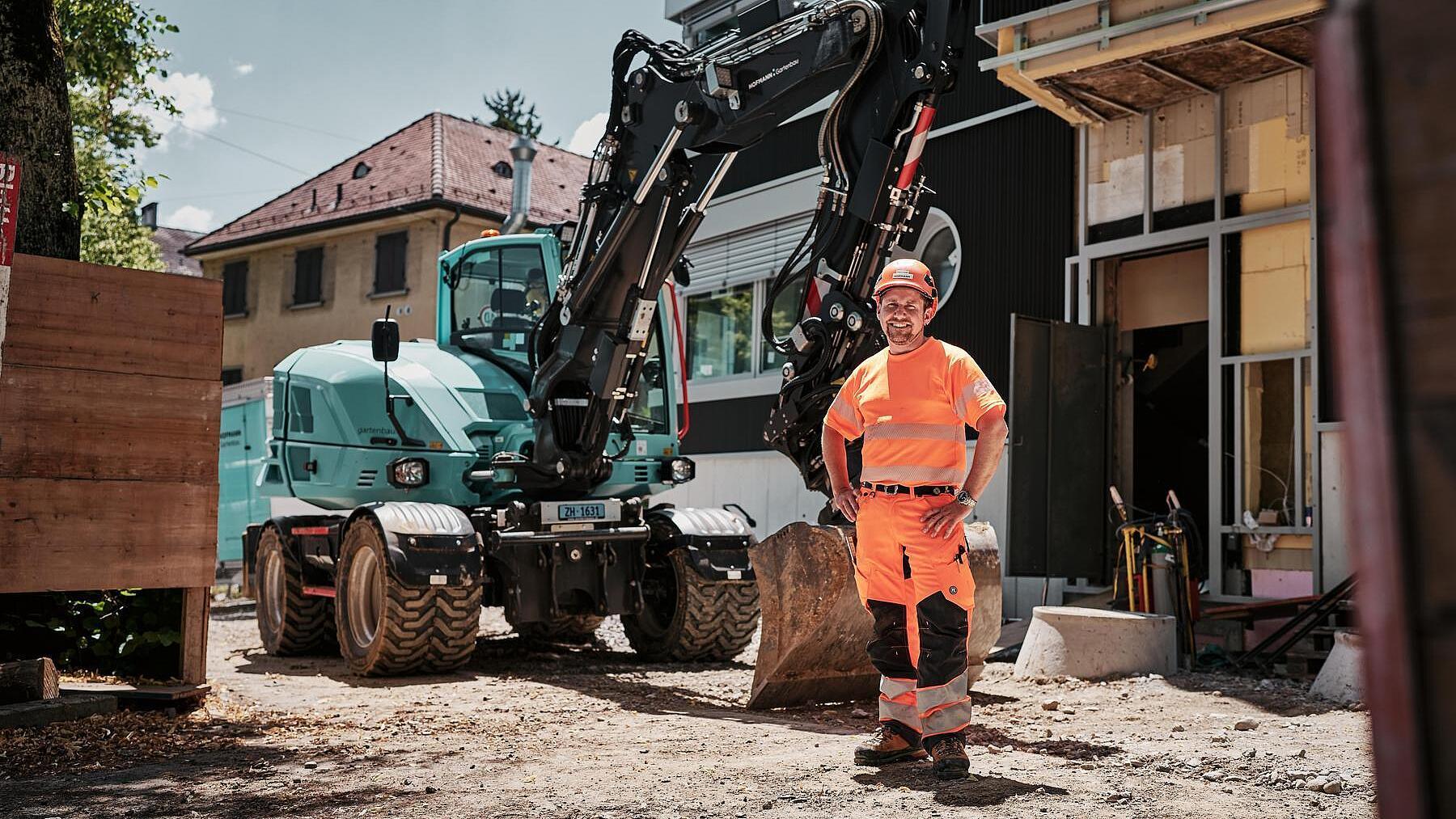 Operator wearing high-visibility workwear standing beside a Wacker Neuson EW100 wheeled excavator at an urban construction site.