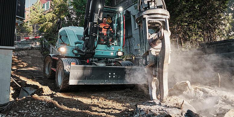 Wacker Neuson EW100 hjulgravemaskin som bruker en hydraulisk hammer til rivningsarbeid i et tettbebygget boligområde.