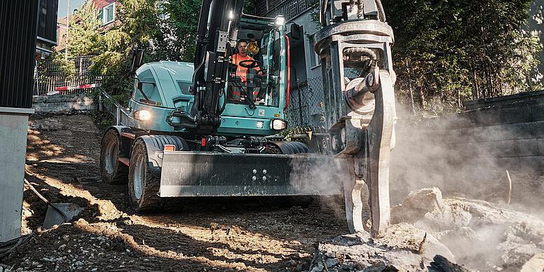 Wacker Neuson EW100 hjulgravemaskine, der anvender en hydraulisk hammer til nedrivningsarbejde i et tætbebygget boligområde.
