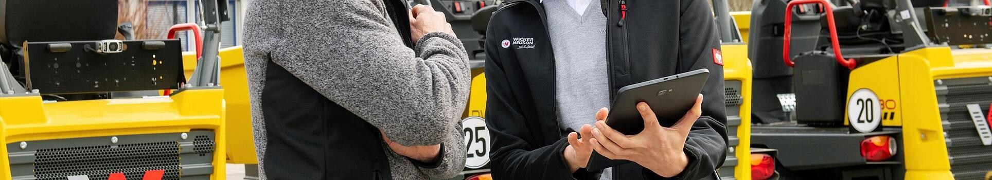 Smiling Wacker Neuson employee with tablet advising smiling customer in front of Wacker Neuson construction machines.