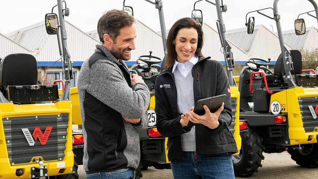 Smiling Wacker Neuson employee with tablet advising smiling customer in front of Wacker Neuson construction machines.