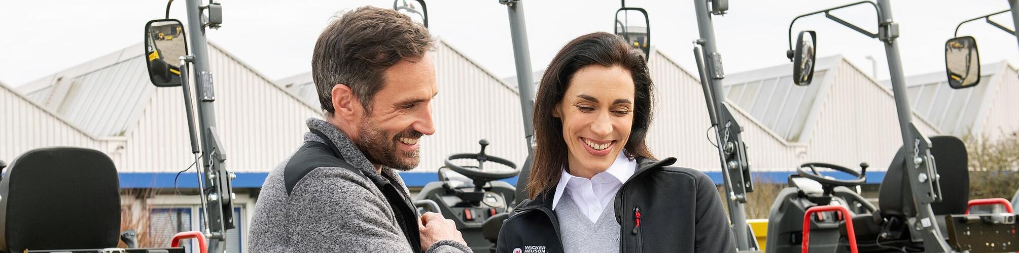 Smiling Wacker Neuson employee with tablet advising smiling customer in front of Wacker Neuson construction machines.