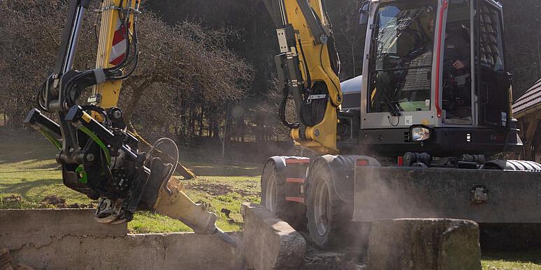 En Wacker Neuson EW100 hjulgravemaskin som bruker en stubbefreser på en skogsklaring, hvor den freser en trestubbe og kaster flis opp i luften, med stablet tømmer og skog i bakgrunnen.