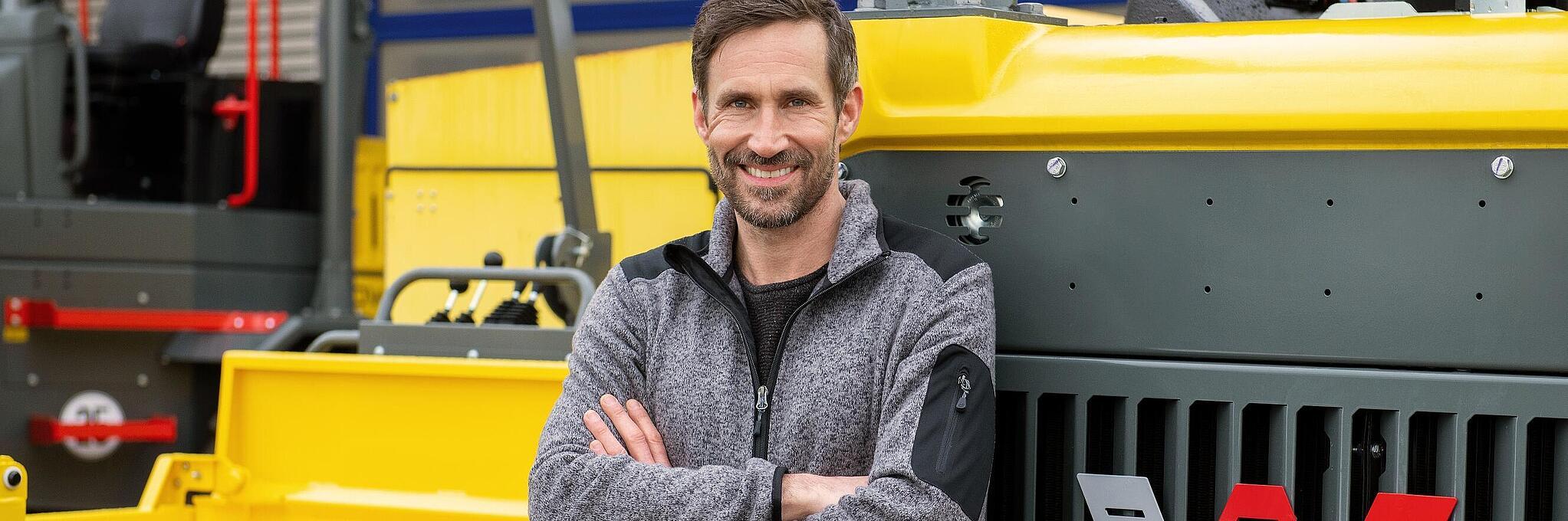 Gülümseyen Wacker Neuson personeli kollarını çapraz bağlamış vaziyette bir Wacker Neuson inşaat makinasına yaslanıyor.