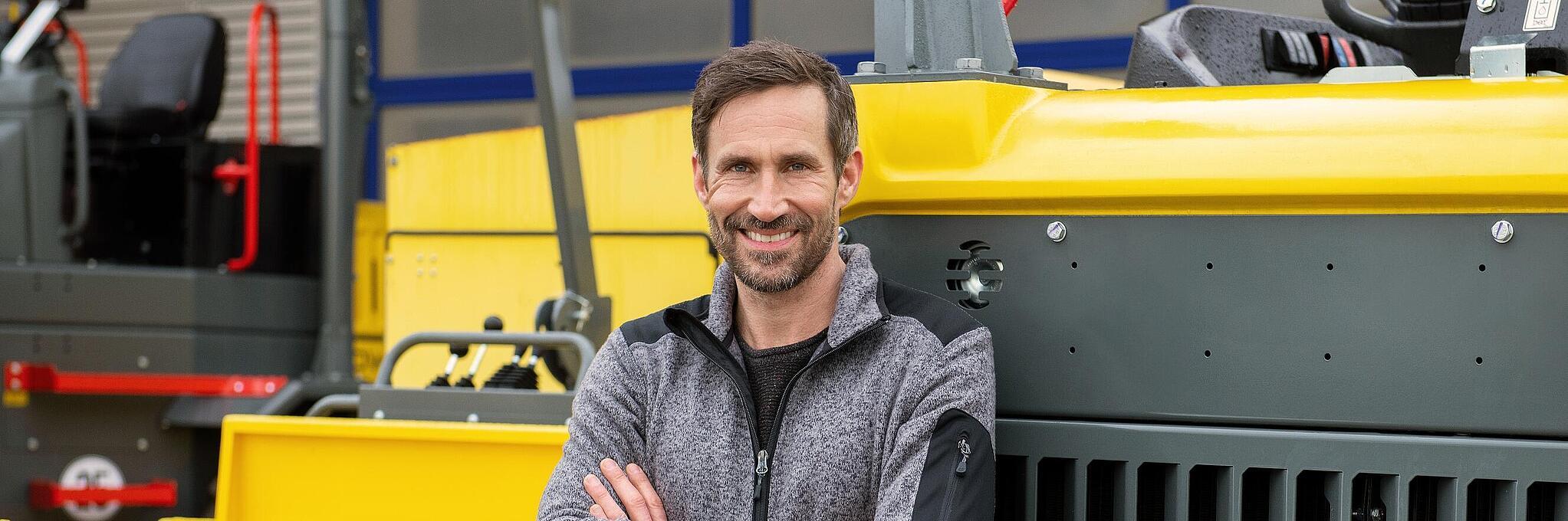 Un collaborateur Wacker Neuson souriant et bras croisés s’appuie sur une machine de construction Wacker Neuson.