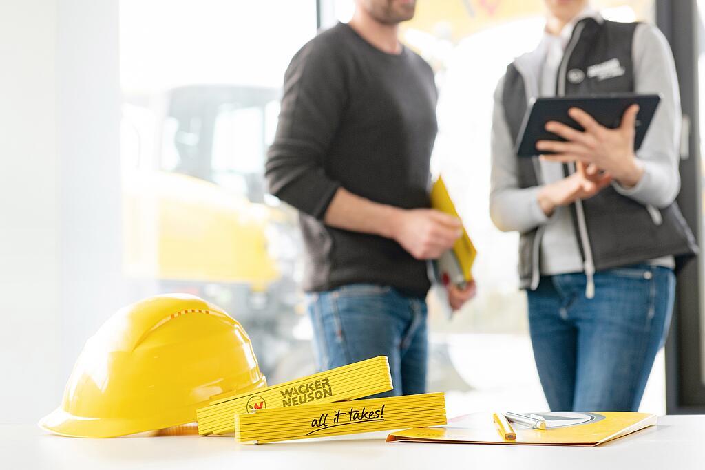 Merchandiseartikler fra Wacker Neuson, der ligger på et bord, i forgrunden, medarbejdere hos Wacker Neuson i baggrunden.
