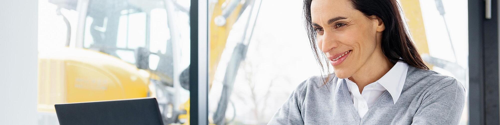 Сотрудница компании Wacker Neuson сидит с улыбкой за ноутбуком.