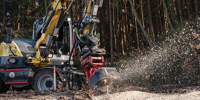 Wacker Neuson EW100 wheeled excavator operating a stump grinder in a forest clearing, grinding a tree stump and throwing wood chips into the air, with stacked logs and woodland in the background.