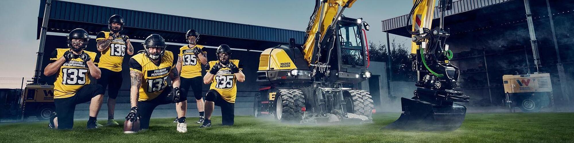 Tiltrotator ek parçası takılı Wacker Neuson EW135 tekerlekli ekskavatör, gece stadyumun projektör ışıkları altında bir Amerikan futbolu takımının yanında konumlandırılmış.