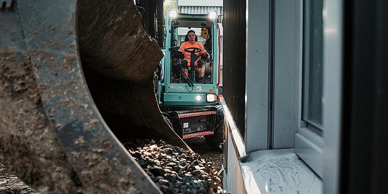 En Wacker Neuson EW100-gravemaskine, der transporterer tilslagsmateriale med skovlen på et trangt byggepladsområde ved siden af en bygning.