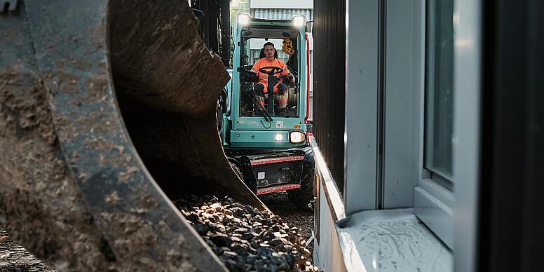 Wacker Neuson EW100 excavator transporting aggregate with a bucket in a narrow construction zone beside a building.