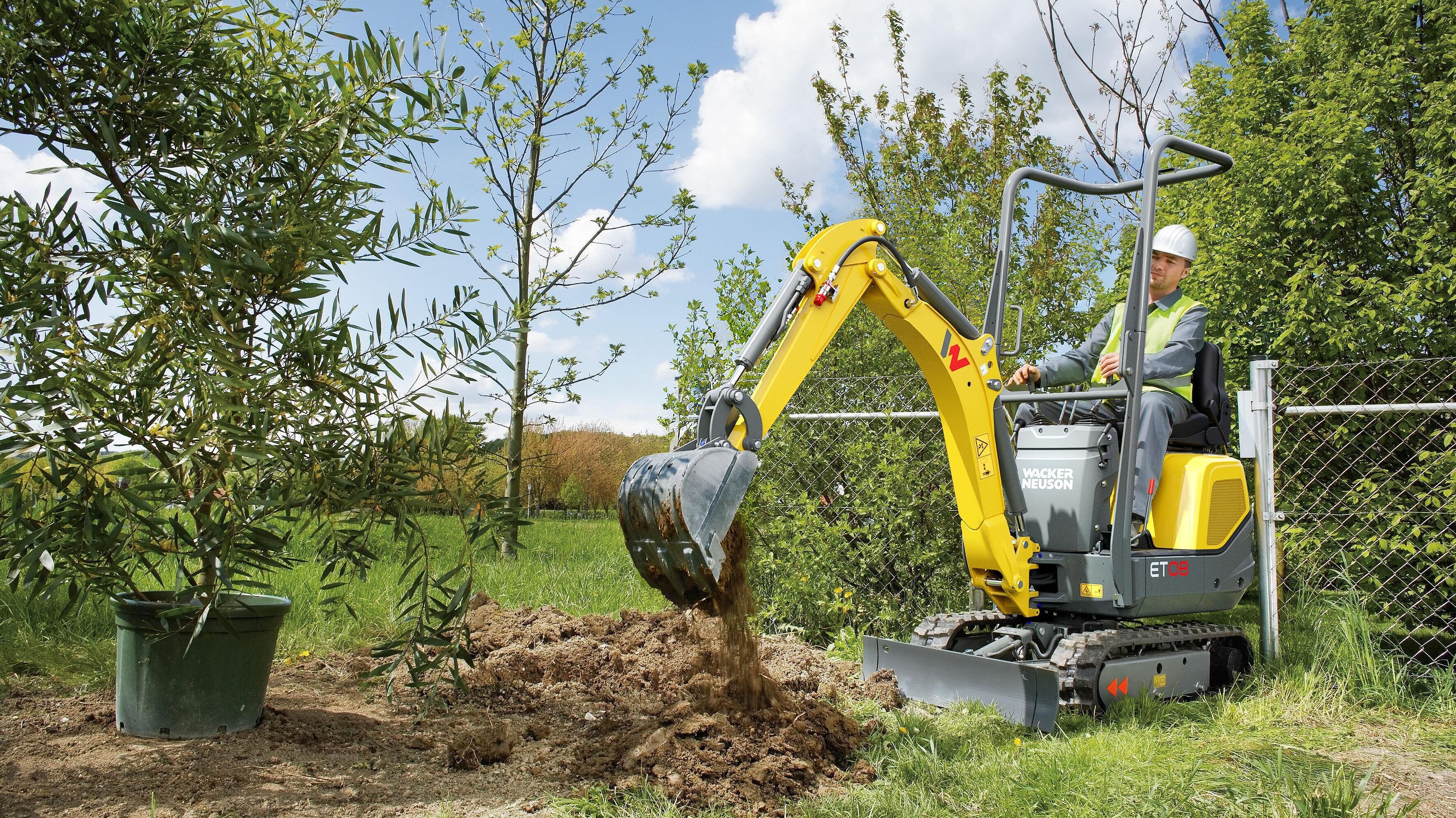 A construction worker wearing a hard hat and high-visibility vest operates a small yellow Wacker Neuson mini excavator. The machine digs up soil to prepare a hole in the garden for planting a tree.