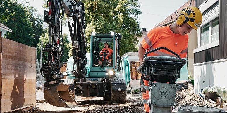 Construction worker operating a Wacker Neuson rammer while the EW100 excavator works in the background.