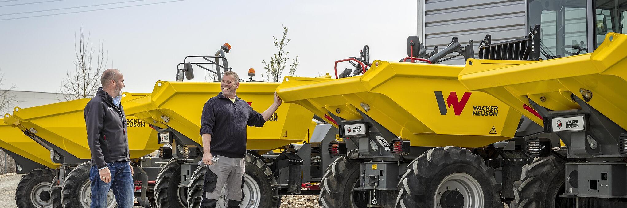 Two men are standing next to several yellow Wacker Neuson dumpers outdoors.