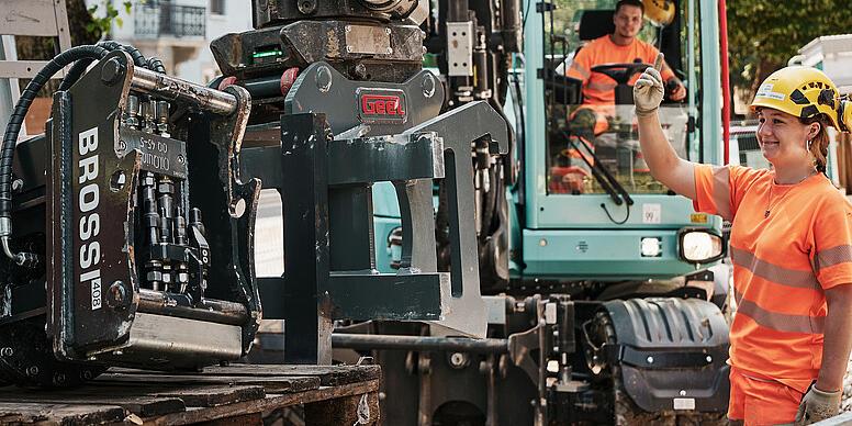 Operator preparing a hydraulic attachment on a Wacker Neuson EW100 wheeled excavator at an urban construction site.