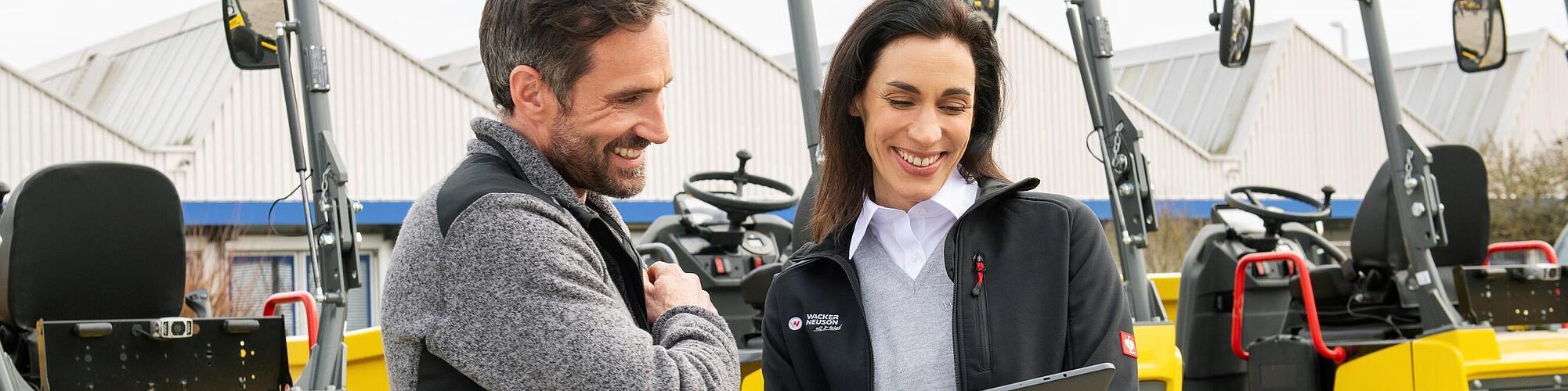 Smilende Wacker Neuson medarbeider med bærbar PC veileder smilende kunde foran Wacker Neuson anleggsmaskiner.