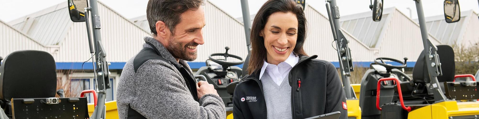 En leende medarbetare från Wacker Neuson håller en surfplatta samtidigt som hon ger råd till en kund framför maskiner från Wacker Neuson.