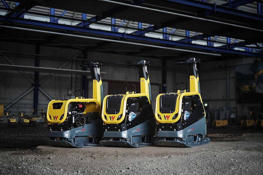Tre gulgrå vibrationsplader fra Wacker Neuson står ved siden af hinanden i en hal på et sandet underlag. Det drejer sig om modellerne BPU 52, DPU 62 og DPU 82. Maskinerne har høje føringshåndtag, kompakt konstruktion og er designet til professionel brug i jordarbejde. I baggrunden kan man se flere maskiner i hallen.