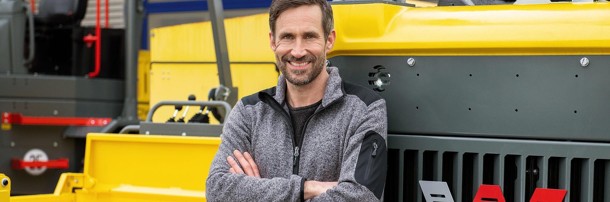 Smiling Wacker Neuson employee, arms crossed, leaning against a Wacker Neuson construction machine.